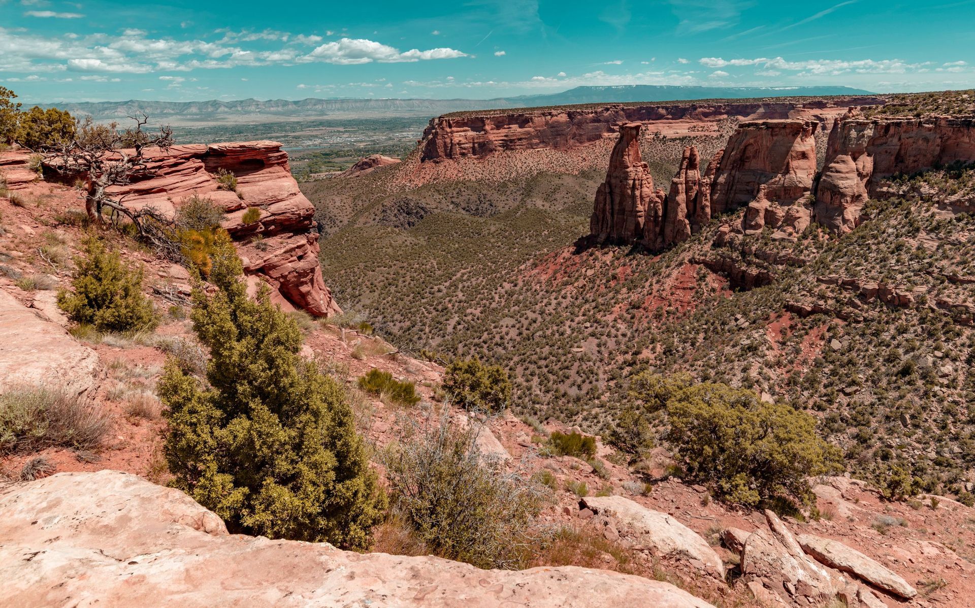 Rocky canyon landscape with red cliffs, sparse shrubs, and a distant green valley under a blue sky