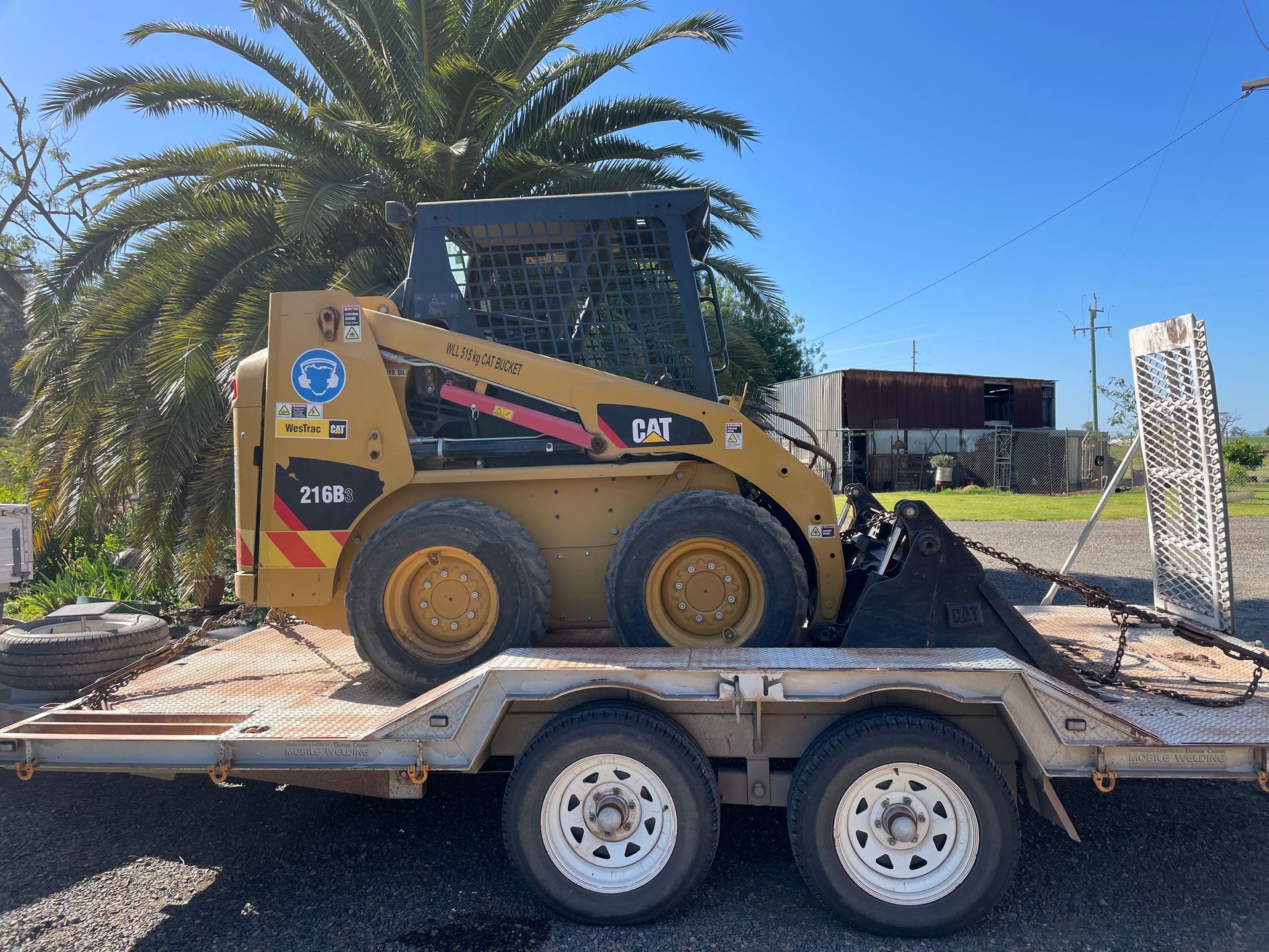 A yellow Caterpillar skid steer loader on a trailer, outdoors on a sunny day.