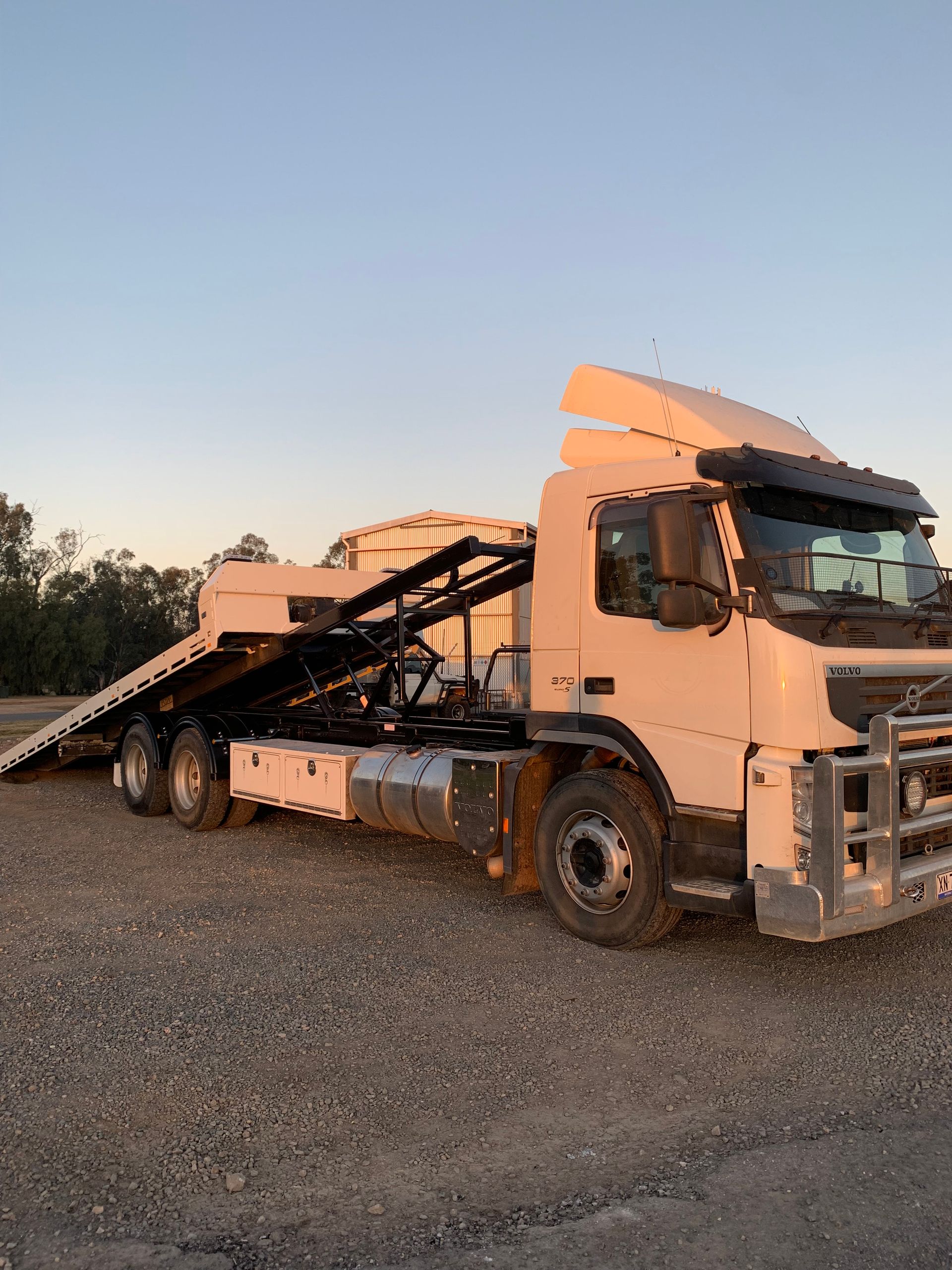 White truck with a flatbed trailer on a gravel lot. It's facing right, with a blue sky in the background.