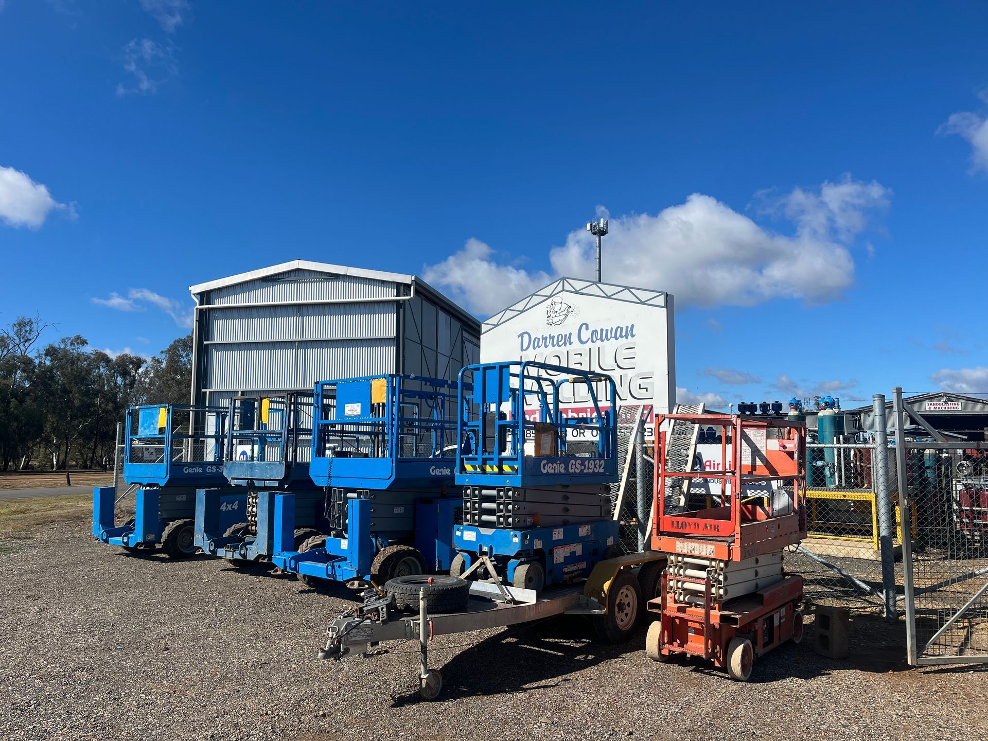 Several aerial lifts of various colors are parked in front of a building with a sign that reads 