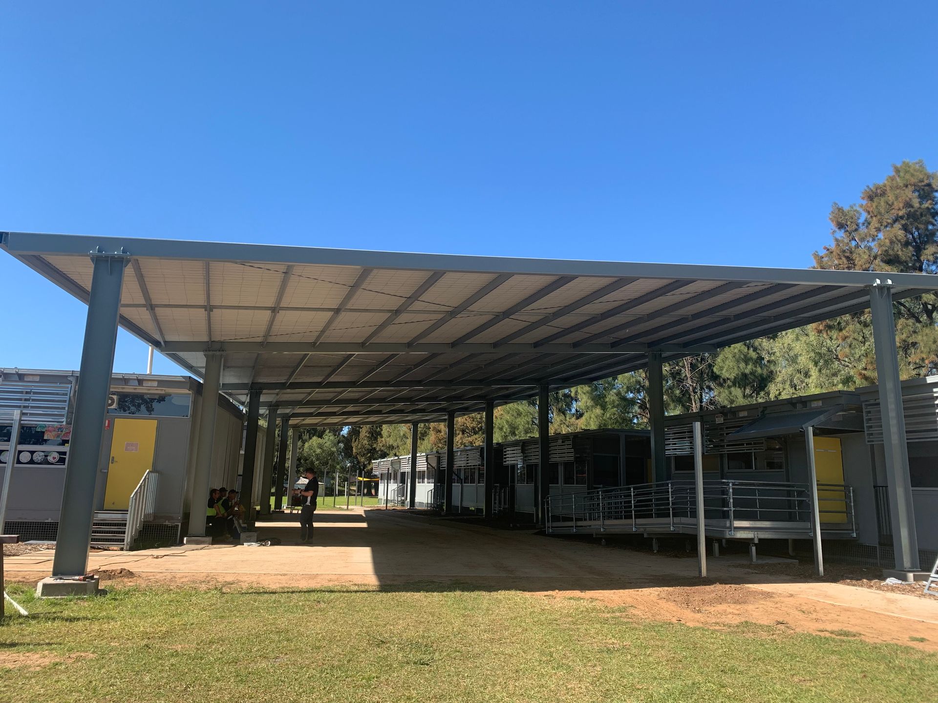 A covered walkway with a metal roof providing shade over several small buildings on a grassy area.