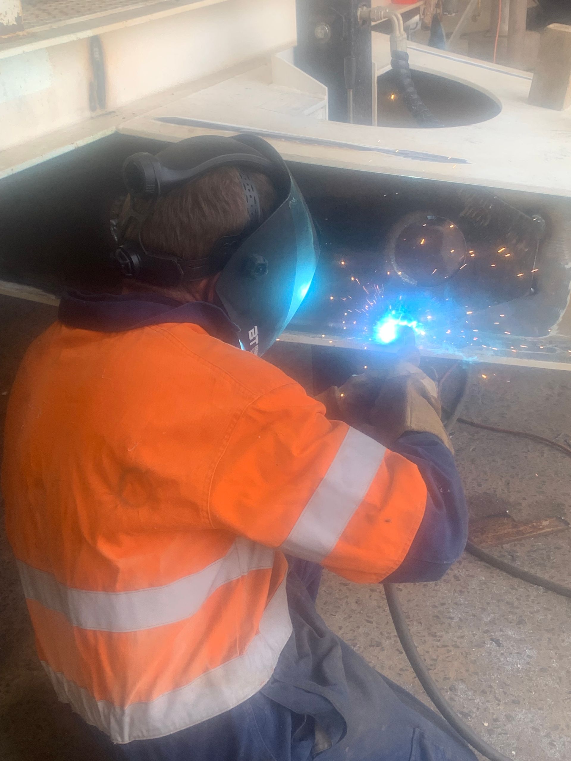 A welder in an orange safety shirt and welding mask uses a torch on a metal structure, producing bright blue light.