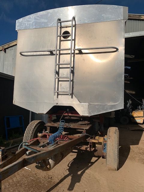 Rear view of an unfinished silver metal trailer with a ladder, attached to a frame with wheels, sitting in a yard.