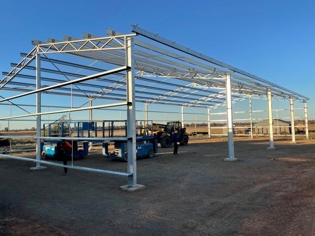 Steel frame of a large barn under construction on a gravel lot, with clear blue sky in background. Two blue lifts are present.