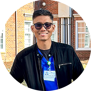 A man wearing sunglasses and a name tag is standing in front of a brick building.