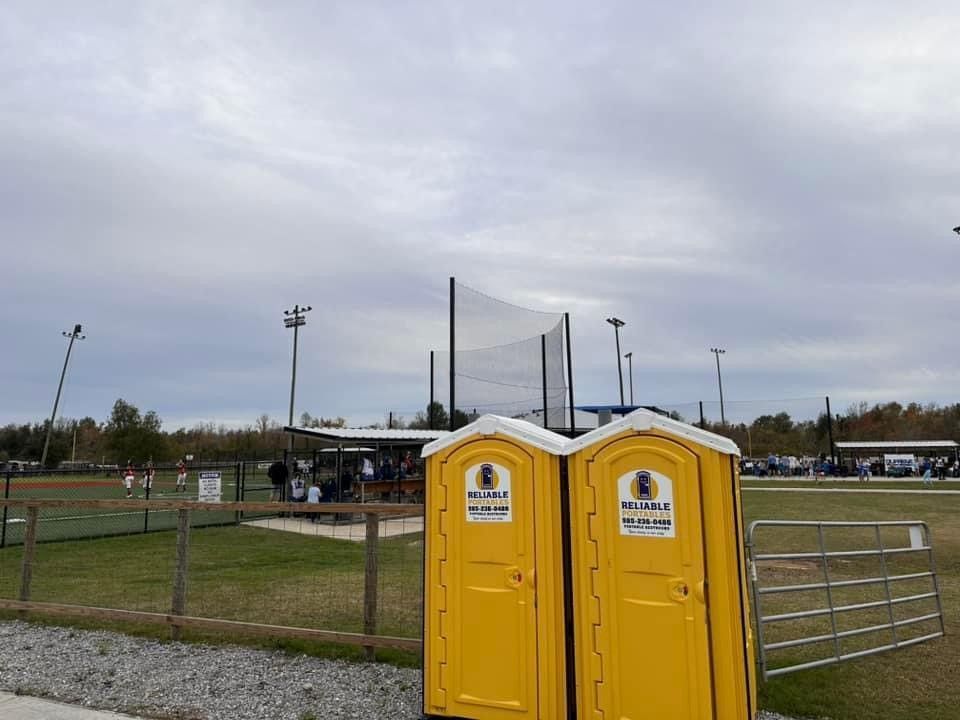 Two yellow portable toilets are sitting next to each other in front of a baseball field.