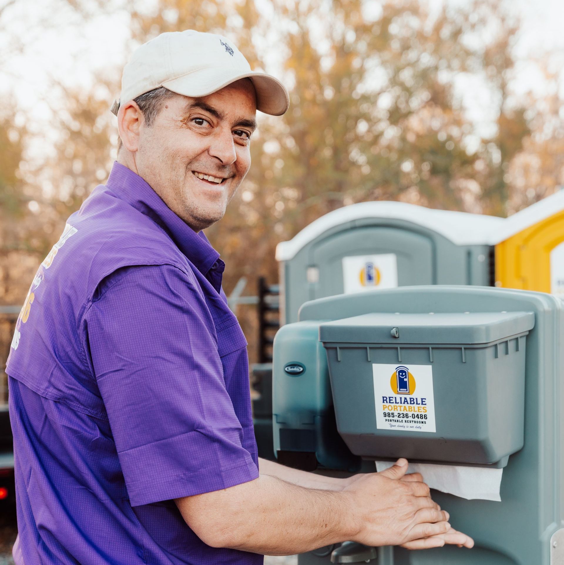 A man in a purple shirt is standing in front of a portable toilet