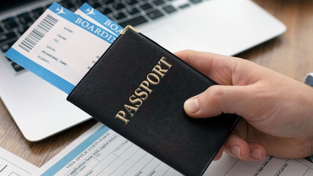 A person is holding a passport and a ticket in front of a laptop.