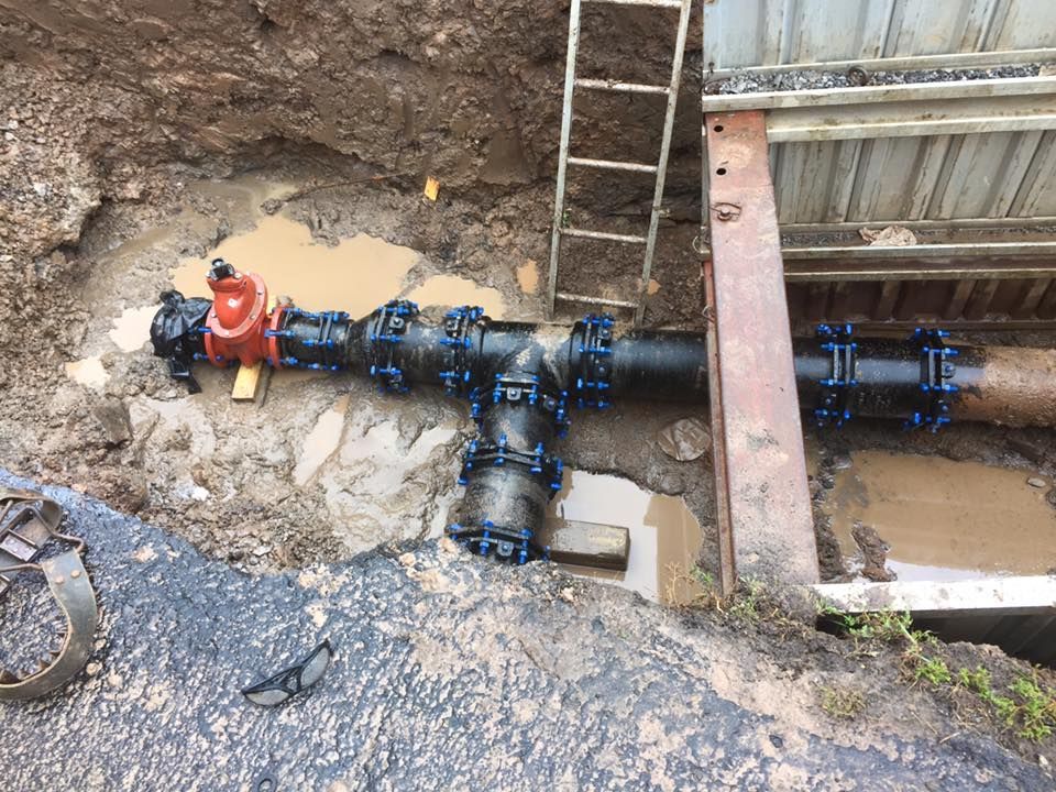 A partially buried black water pipe with a red valve, connected with black fittings, in a muddy excavation.
