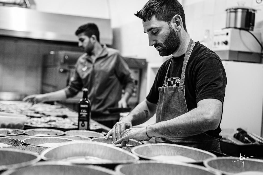 Due chef in cucina preparano il cibo su un bancone con numerose teglie. Uno è concentrato sul suo compito, l'altro lavora dietro le quinte.