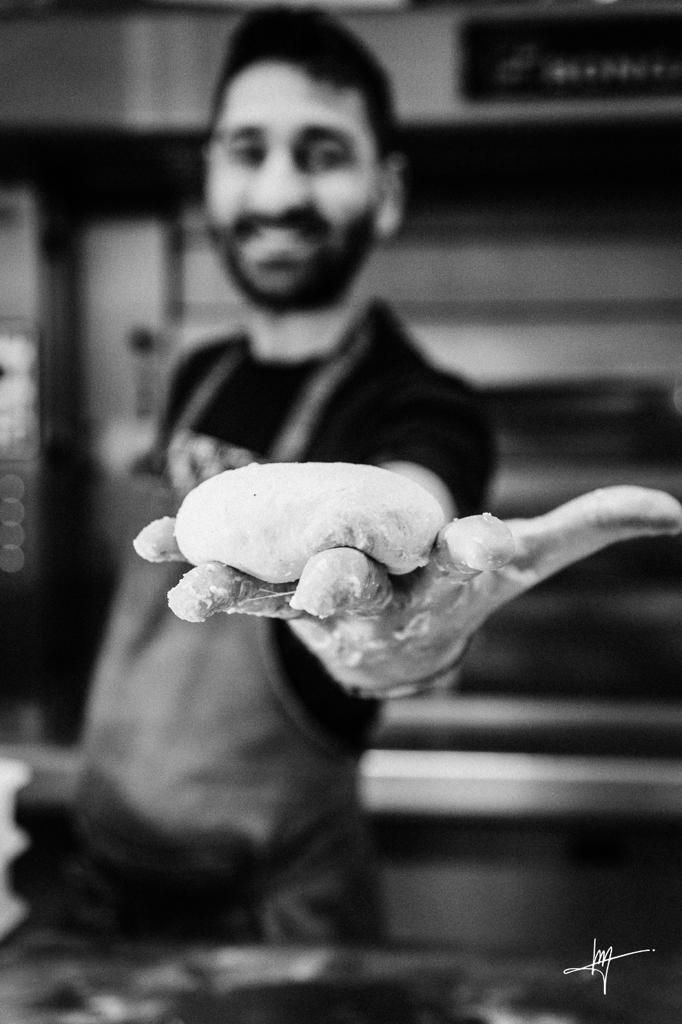 Un fornaio sorridente porge una palla di impasto, allungando il braccio verso l'osservatore. Foto in bianco e nero, ambientata in una cucina.
