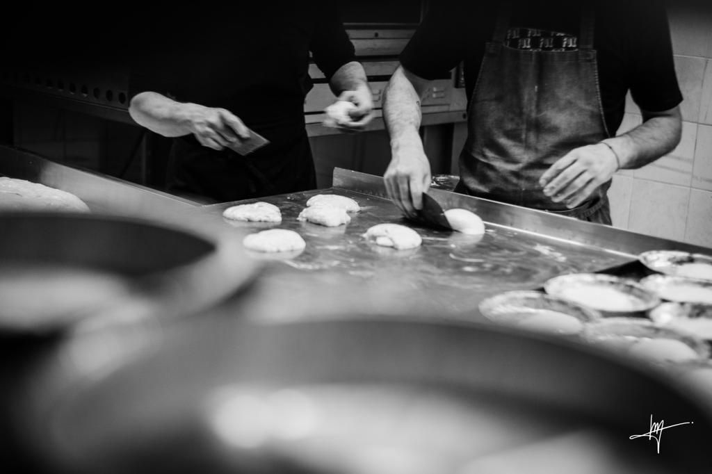 Due persone preparano il cibo in una cucina commerciale. Una stende l'impasto, l'altra dispone gli ingredienti su una piastra, probabilmente preparando prodotti da forno.