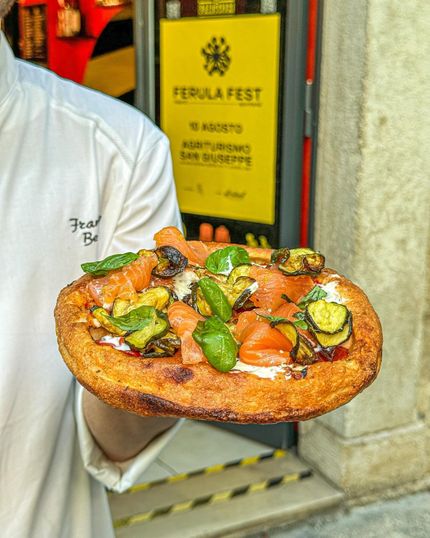Chef che tiene in mano una pizza con salmone, zucchine e basilico davanti a un negozio con un cartello giallo per "Ferula Fest".