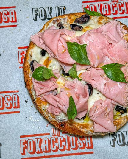 Una pizza rotonda con prosciutto cotto a fette sottili, olive e basilico. Appoggiata su carta con il logo "FOKACCIOSA".