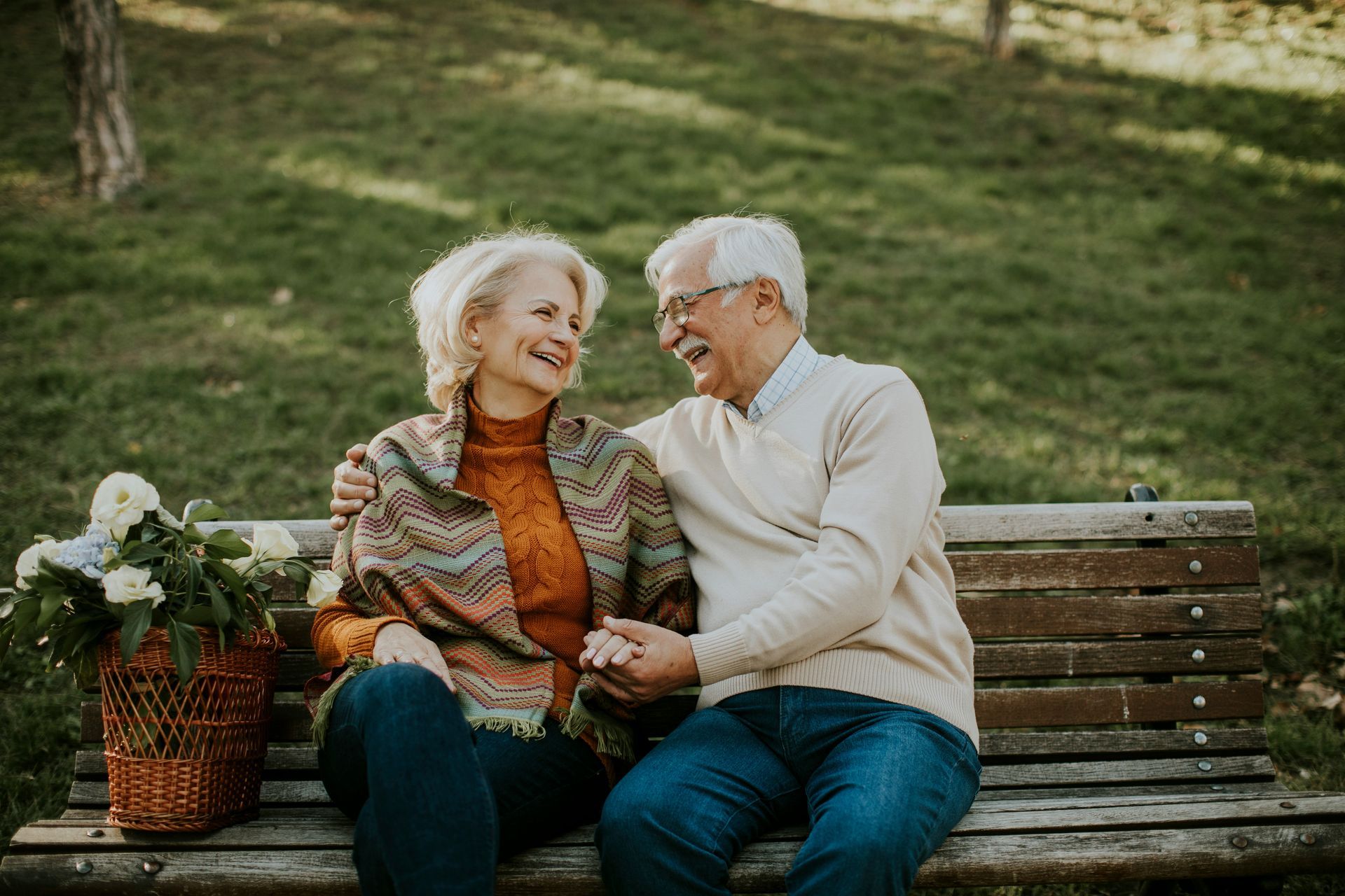 Two people sitting on a park bench, smiling and holding hands beside a basket of flowers.