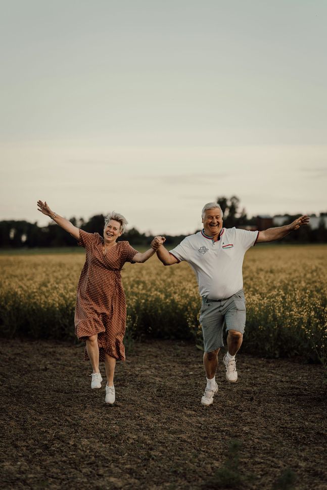 Two people holding hands and jumping through a field at sunset, smiling with arms outstretched.