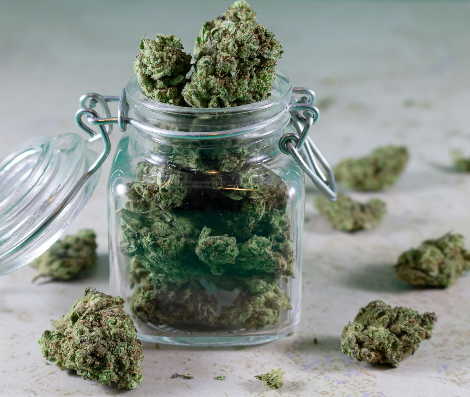 Jar filled with green cannabis buds; some buds scattered around the jar on a light gray surface.
