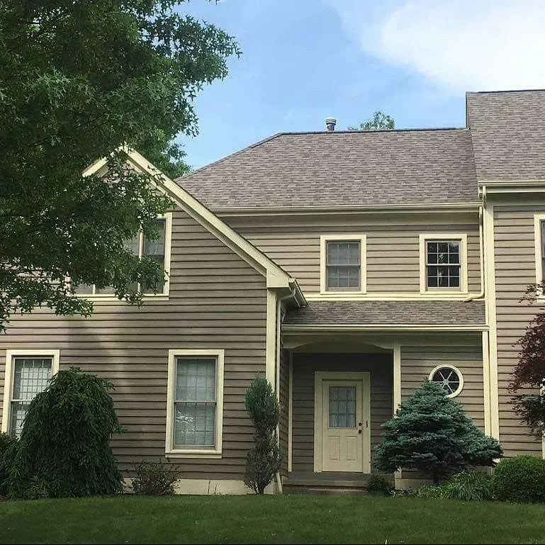 Brown house with tan trim, windows, and door, surrounded by greenery and a blue sky.