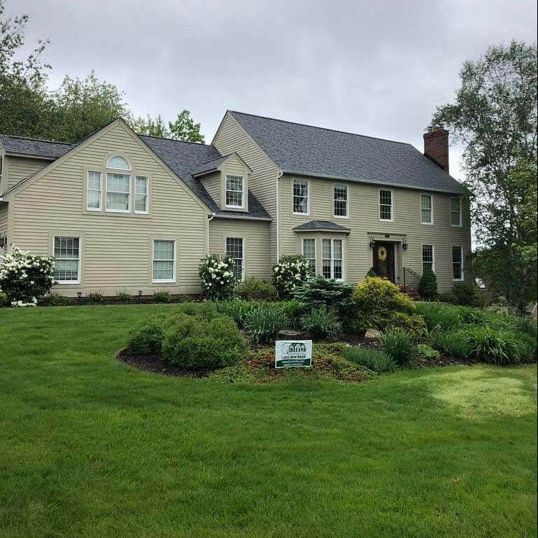 Large beige house with dark roof, manicured lawn, and flower bed.