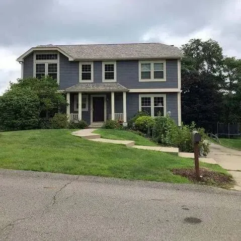 Two-story blue house with porch, lawn, and walkway. Trees and a cloudy sky in the background.