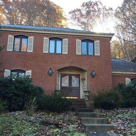 Two-story red brick house with gray roof and shutters. Front door with steps leading up to it. Bushes in front.