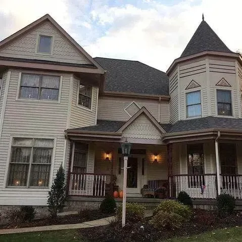 Beige Victorian-style house with a wraparound porch and a turret under a cloudy sky.