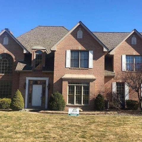 Two-story brick house with a brown roof and white shutters. Front lawn has bushes and dry grass.