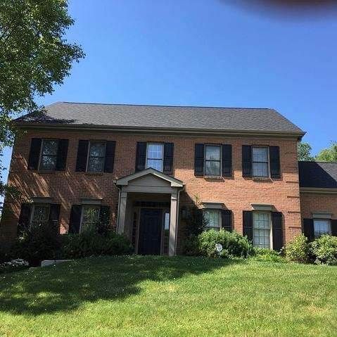 Two-story brick house with black shutters, black roof, and front lawn on a sunny day.