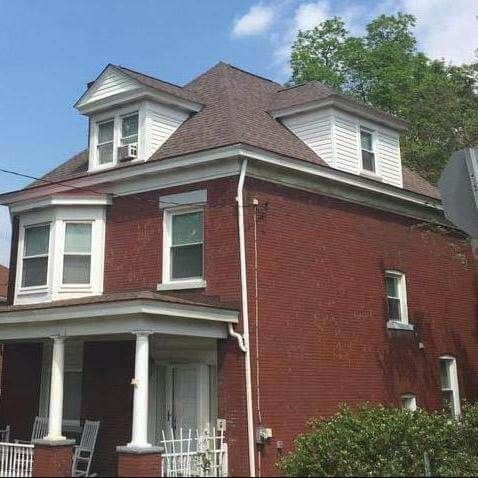Red brick two-story house with white trim, porch, and dormers. Brown roof and clear blue sky.