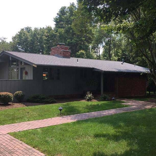 A one-story house with gray siding, brick chimney, and a brick walkway, set in a grassy yard.