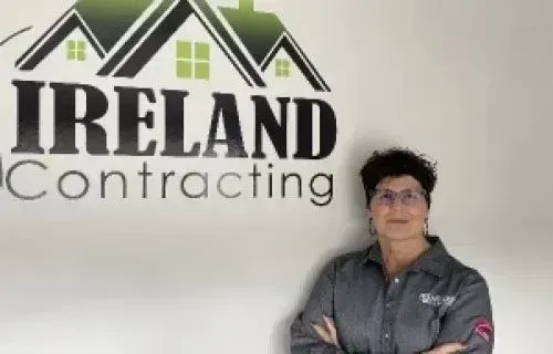 Woman in a gray work shirt stands in front of a company logo that says 