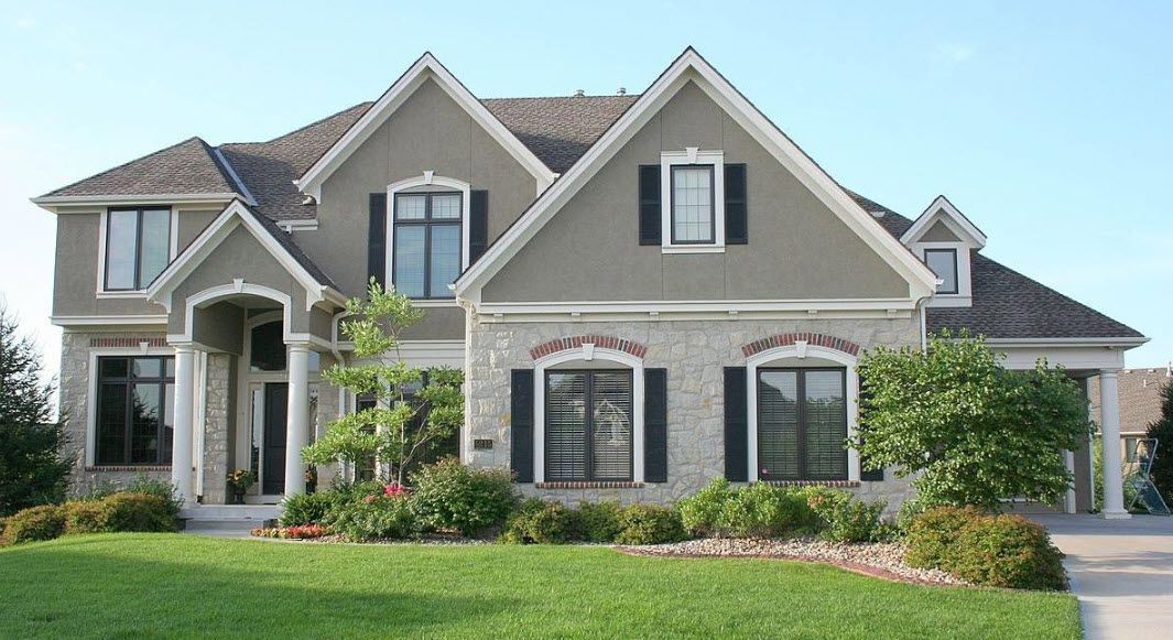 Two-story house with tan siding, brick facade, and black shutters, on a green lawn, under a blue sky.