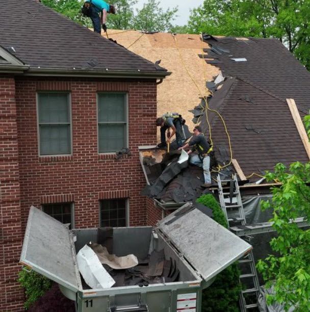 Roofers removing old shingles from a two-story brick house, discarding them into a dumpster below.