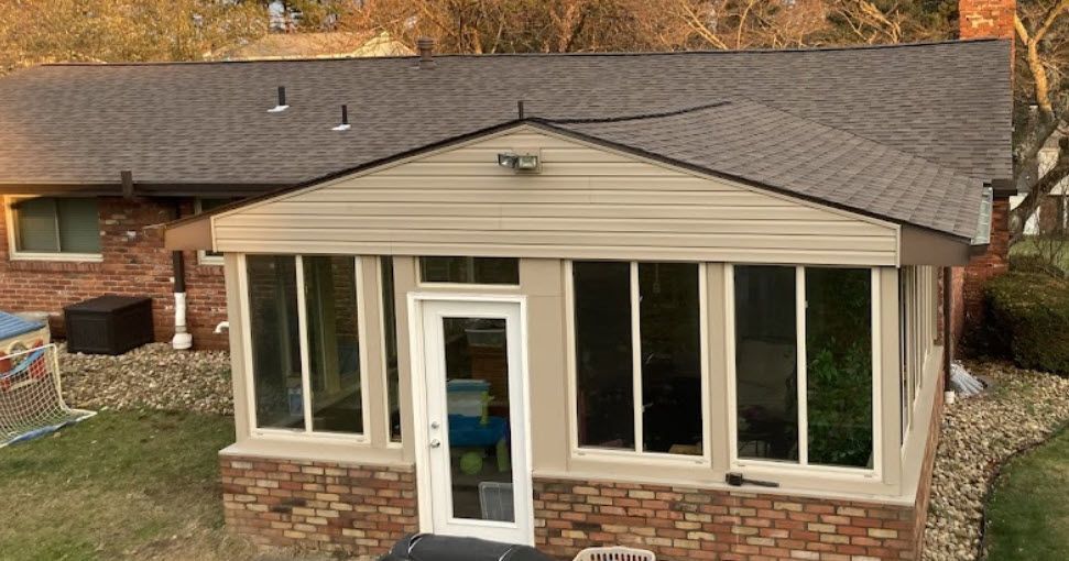 Sunroom addition with brick base, tan siding, large windows, and a glass door; attached to a brick house.