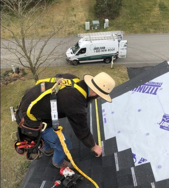 Roofer in safety harness measures shingles on a roof. A white van with roofing logo is in the background.