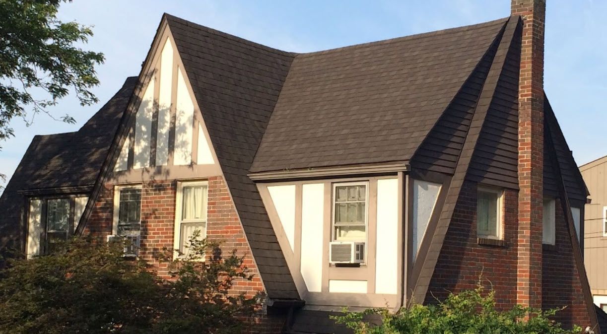 Tudor-style house with brown roof and brick chimney. White and brown facade, windows, and greenery.