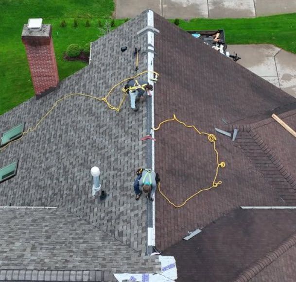 Workers on a roof with gray and brown shingles; safety ropes attached.