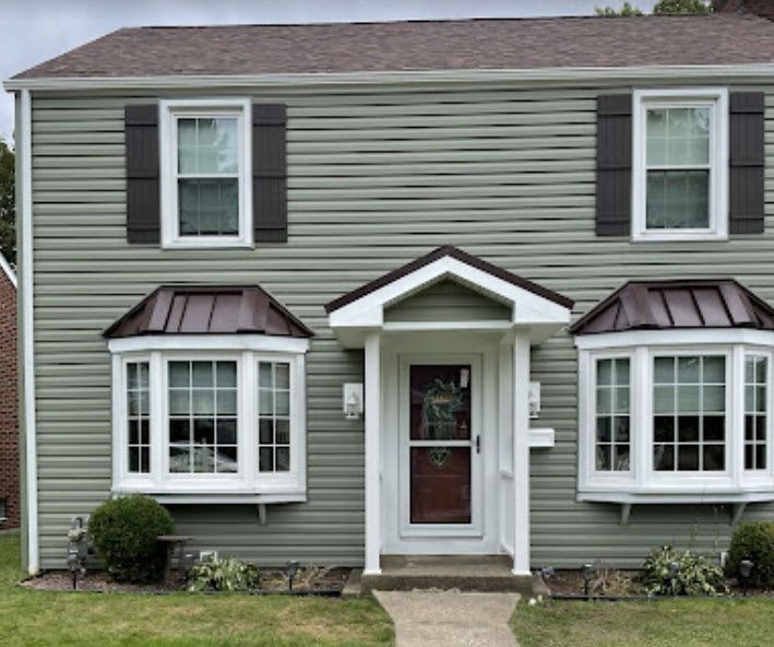 Two-story house with green siding, brown roof and trim, and white-framed windows; two bay windows, door with portico.