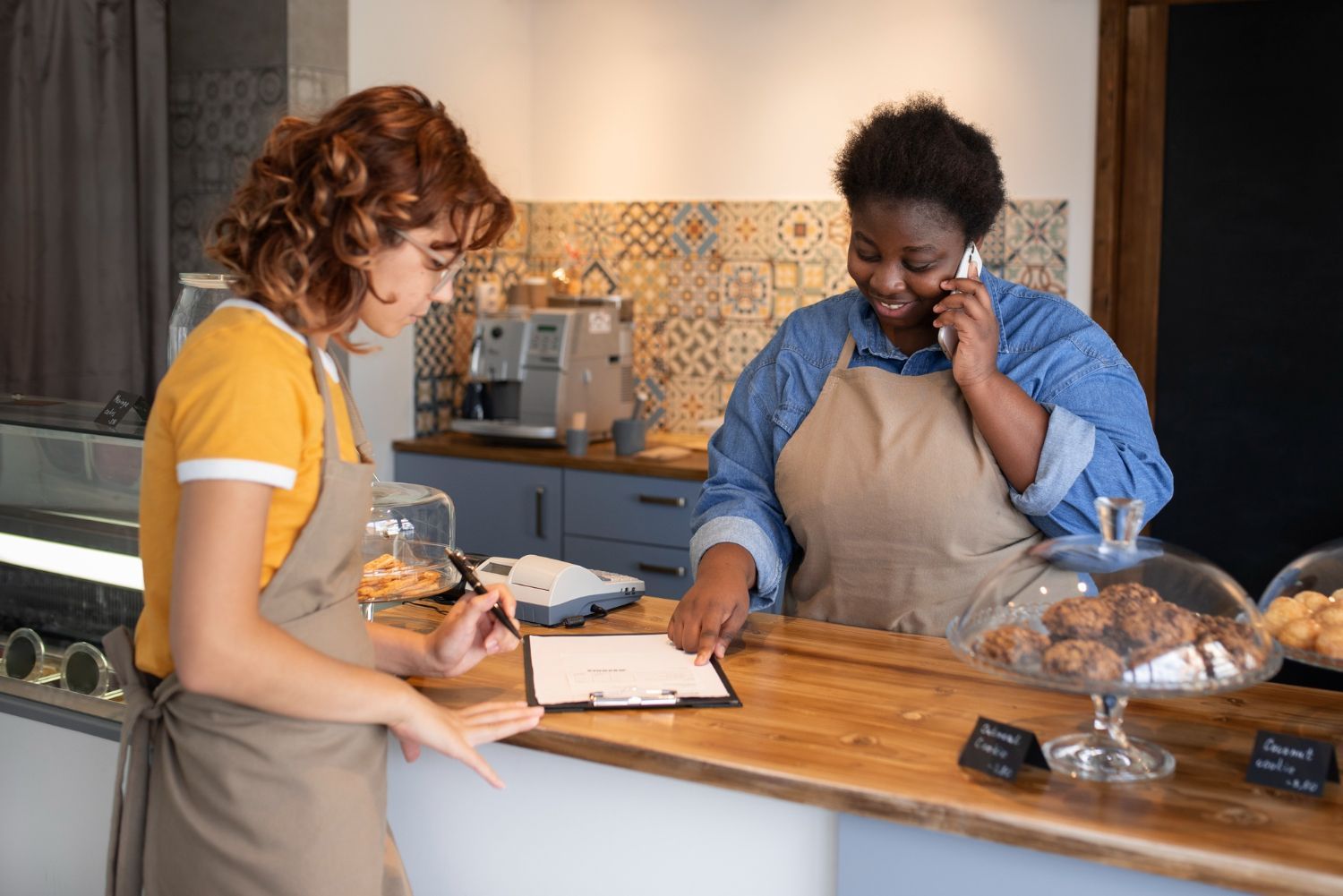 Two people behind a bakery counter, one handing over a pastry in a warm kitchen.