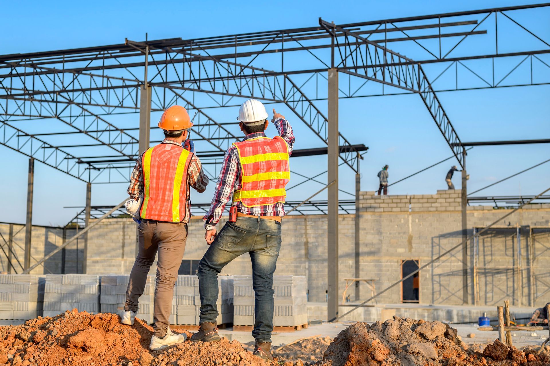 Two construction workers in hard hats and safety vests on a building site with steel framing in the background