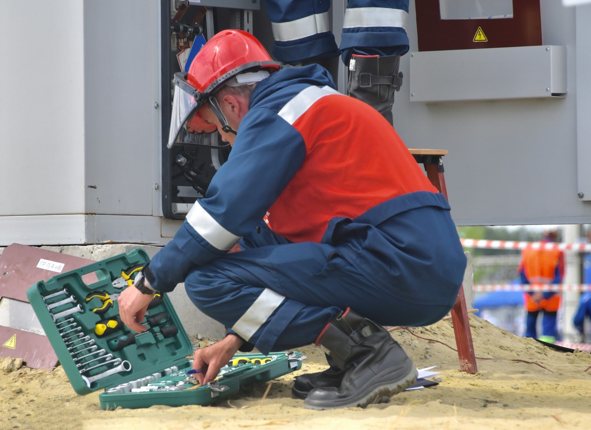 Worker in red helmet kneels by an open utility box, using tools on cables at a construction site.
