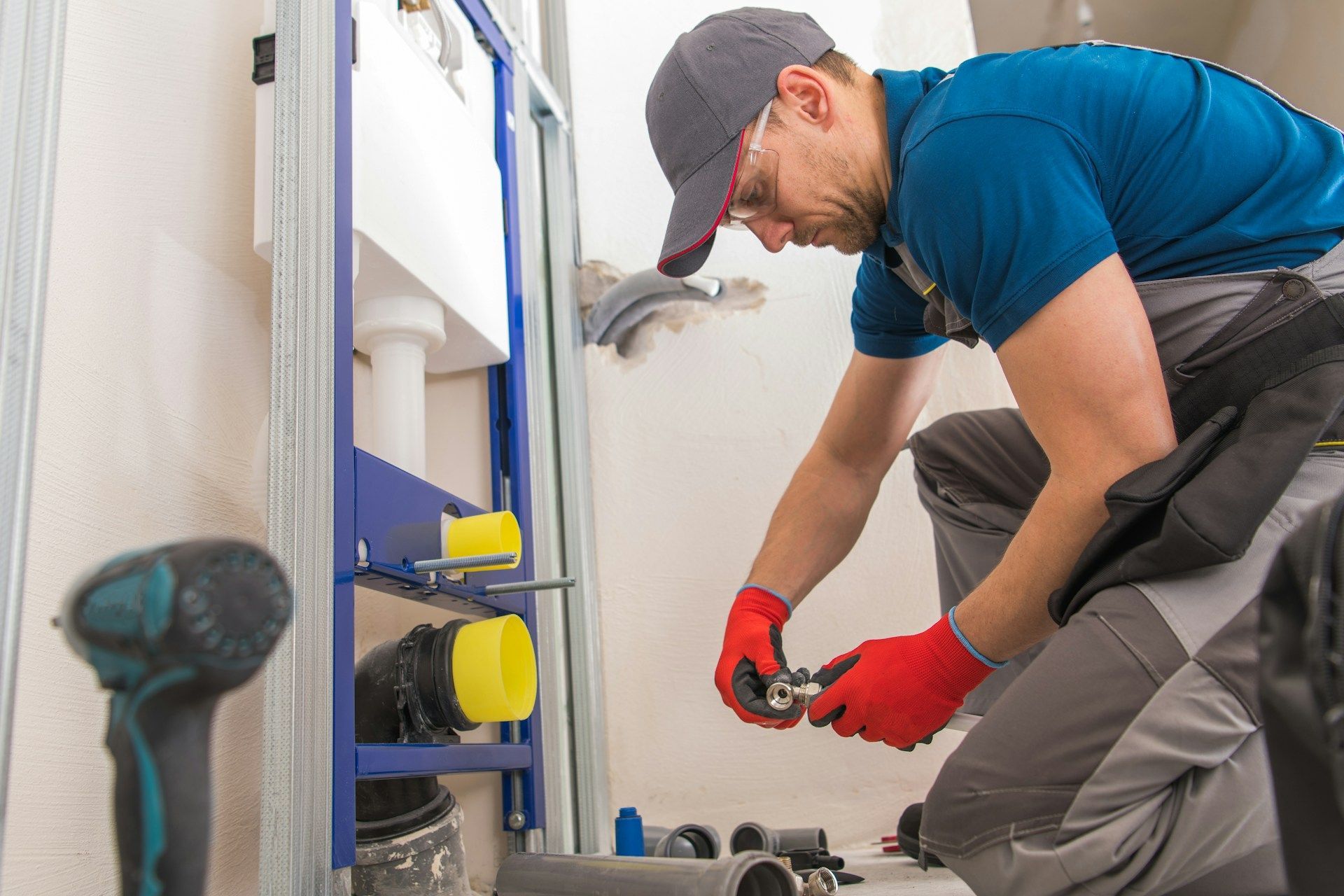 Technician in blue shirt and red gloves repairs plumbing under a sink in a bathroom.