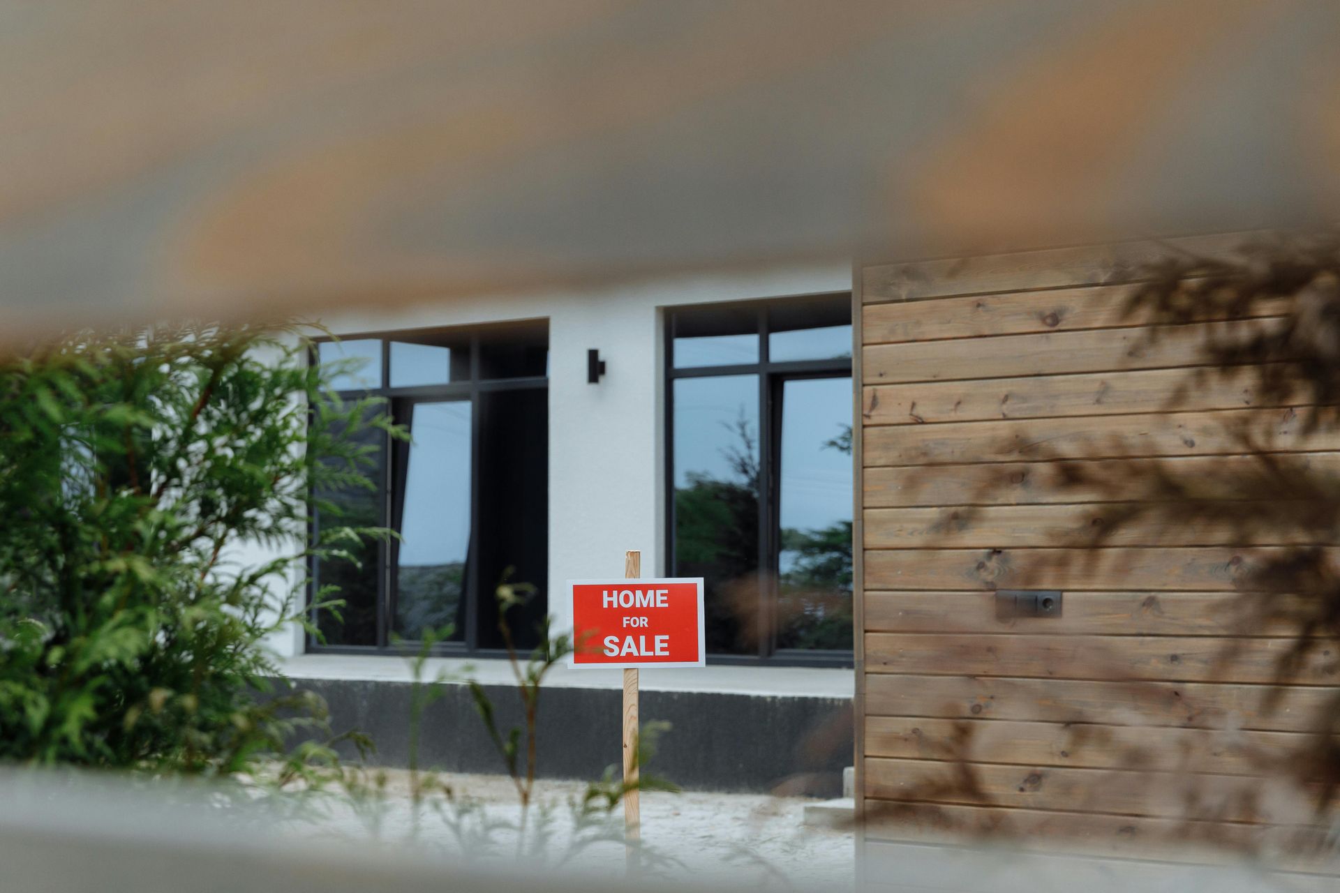House exterior with a red “For Sale” sign in the window and a wooden wall on the right.