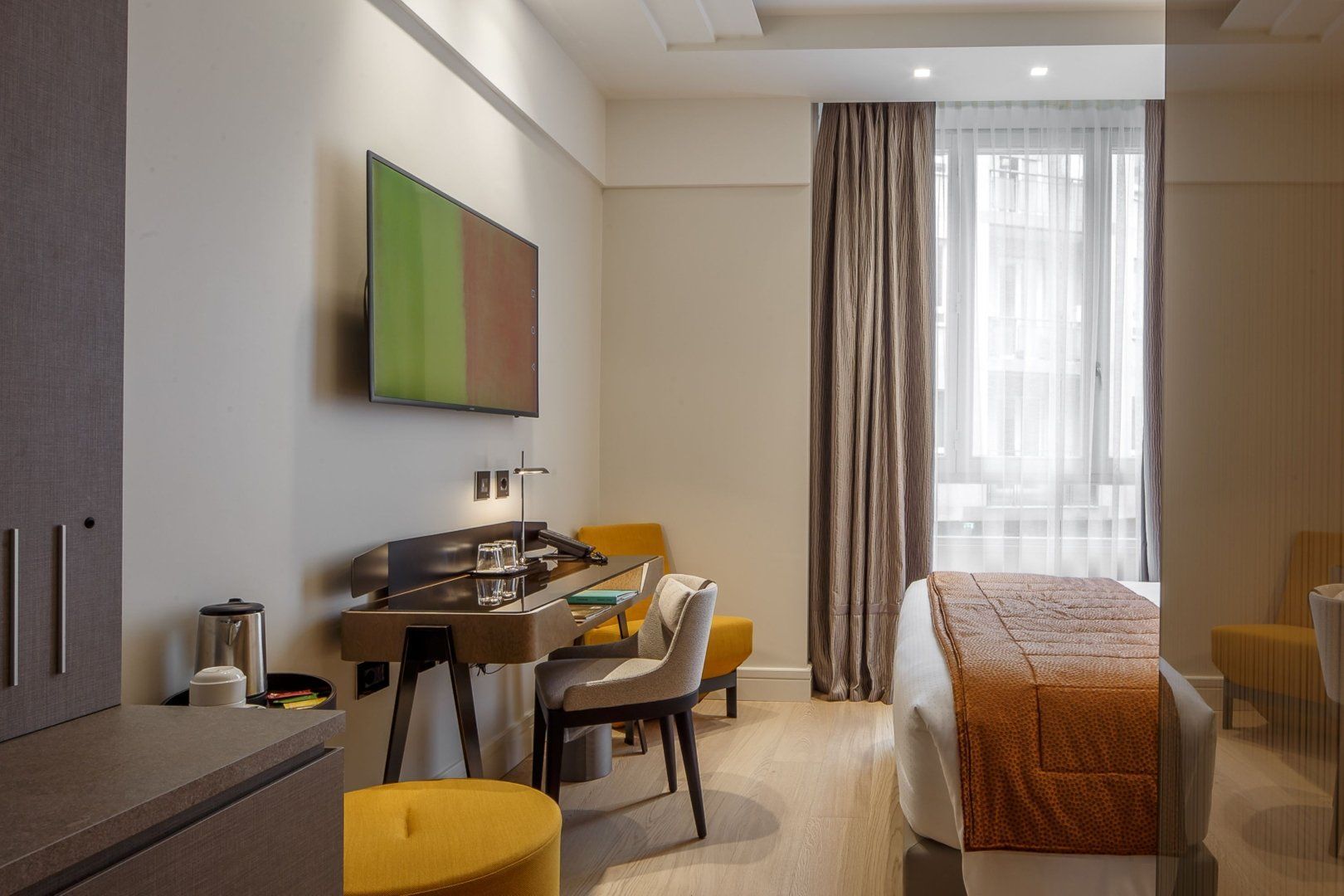 ベッド、木製の机と椅子、黄色のスツール、壁に取り付けられた薄型テレビを備えたモダンなホテルの客室。