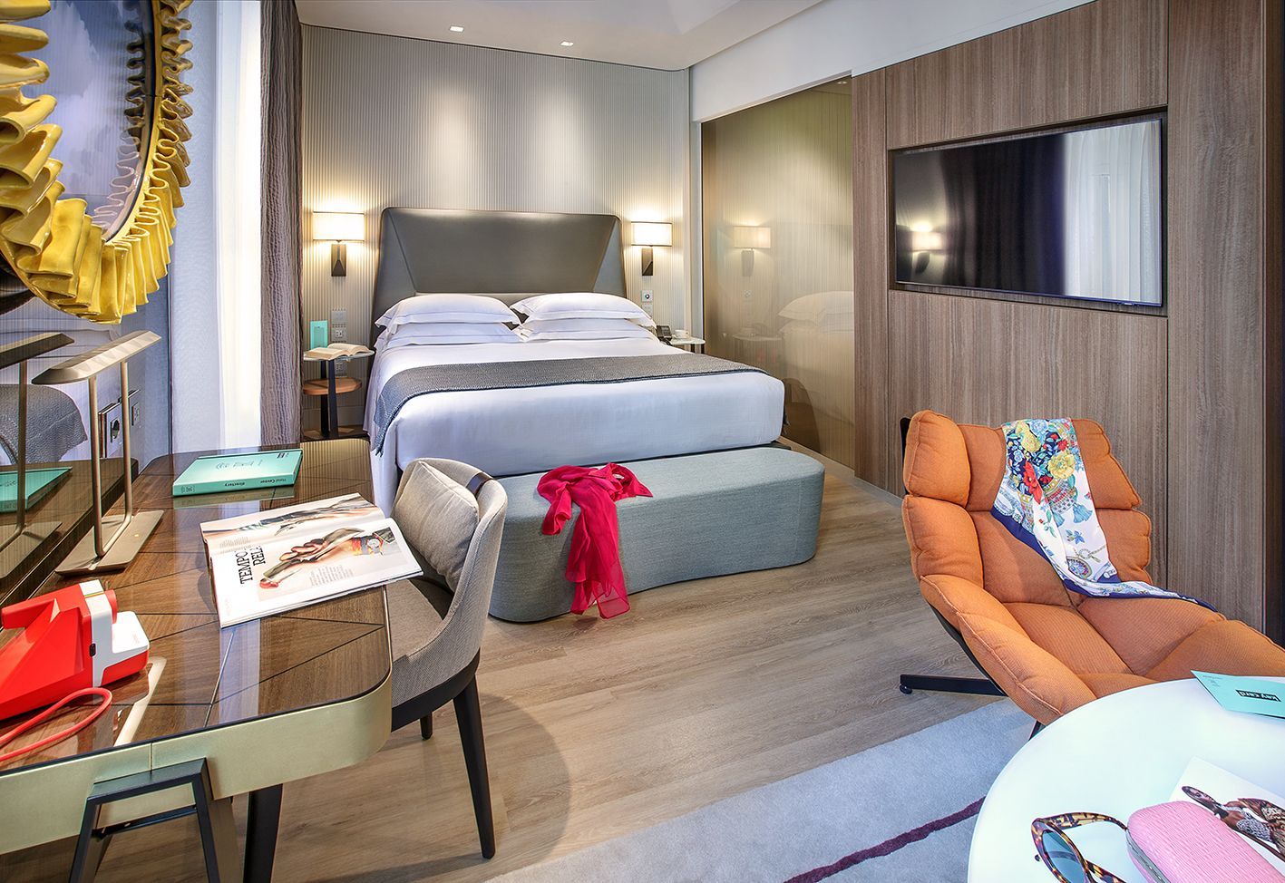A modern hotel room with a neatly made bed, an orange armchair, a wooden desk, and a large decorative circular gold mirror.