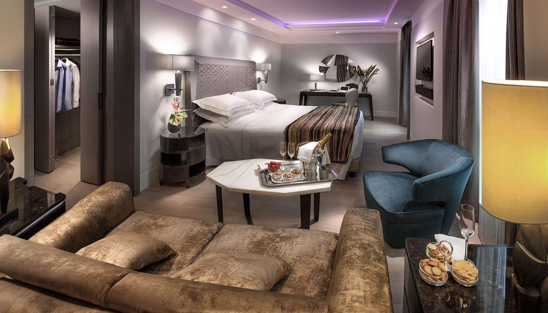 豪华酒店套房，配有特大号床、天鹅绒沙发、蓝绿色扶手椅和摆放着饮料的小餐桌。