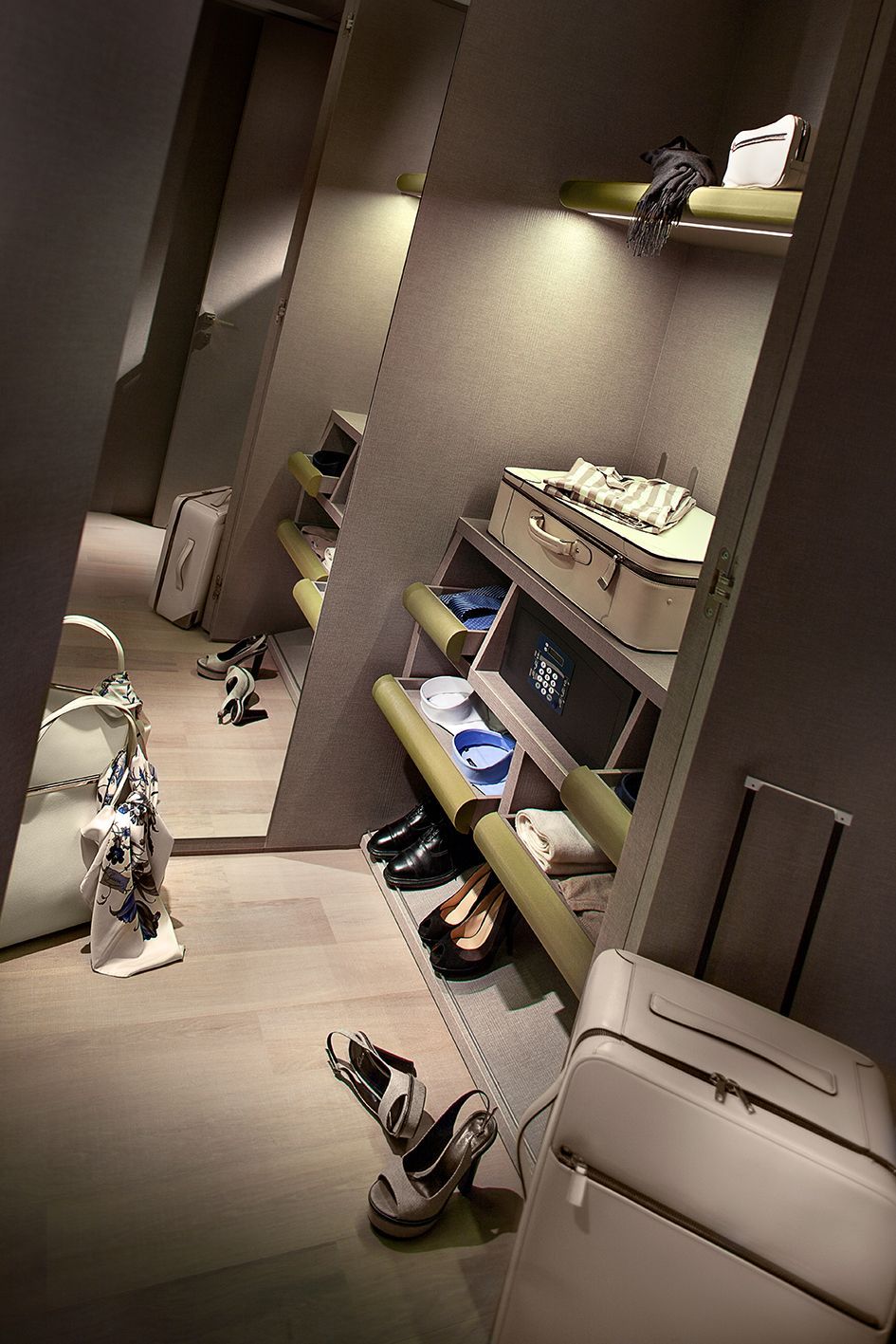 A walk-in closet with shelves holding handbags and shoes, featuring a large mirror and rolling luggage cases on the floor.
