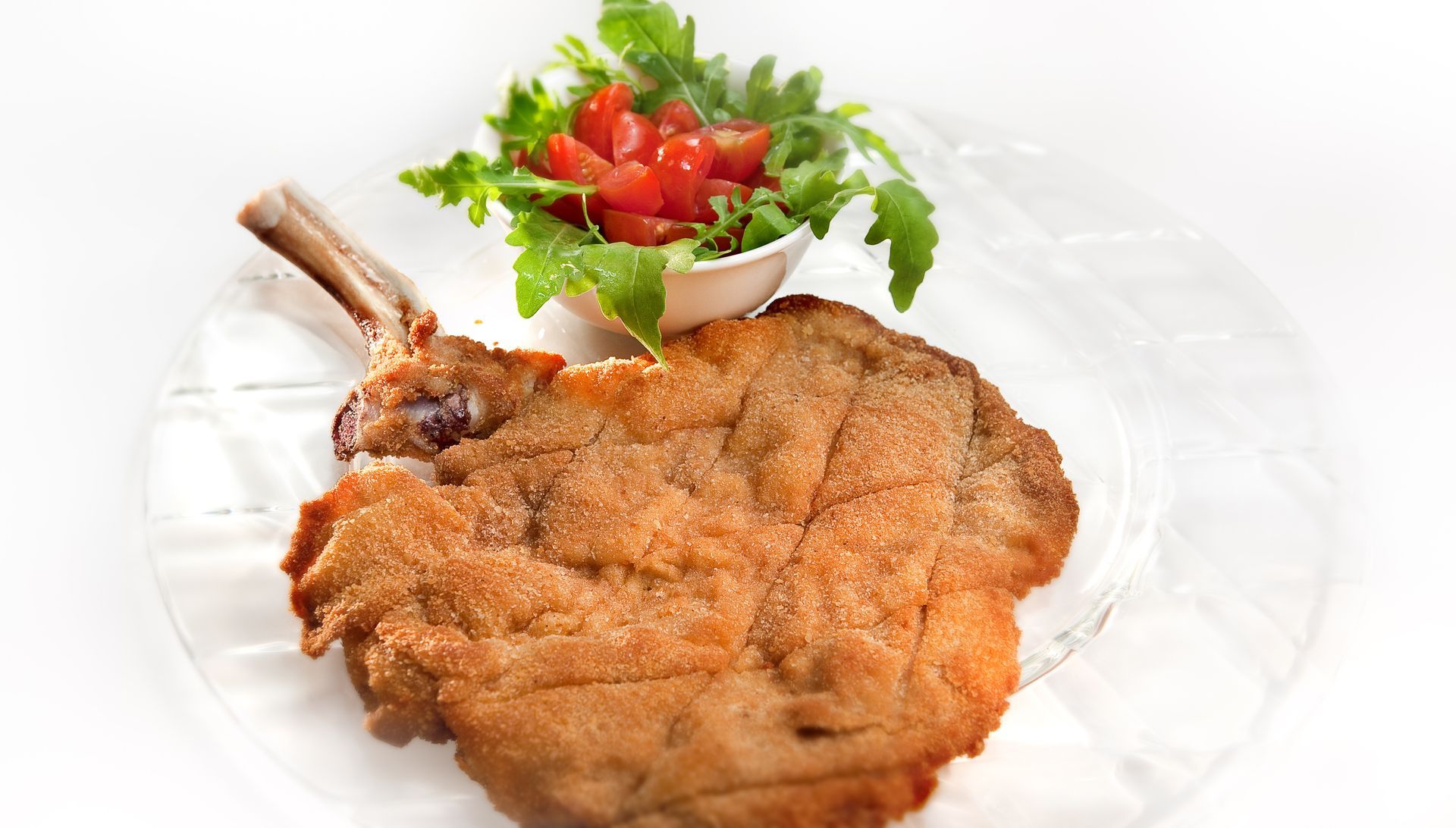 Una costoletta di vitello impanata servita con una ciotolina di insalata fresca di rucola e pomodorini su sfondo bianco.