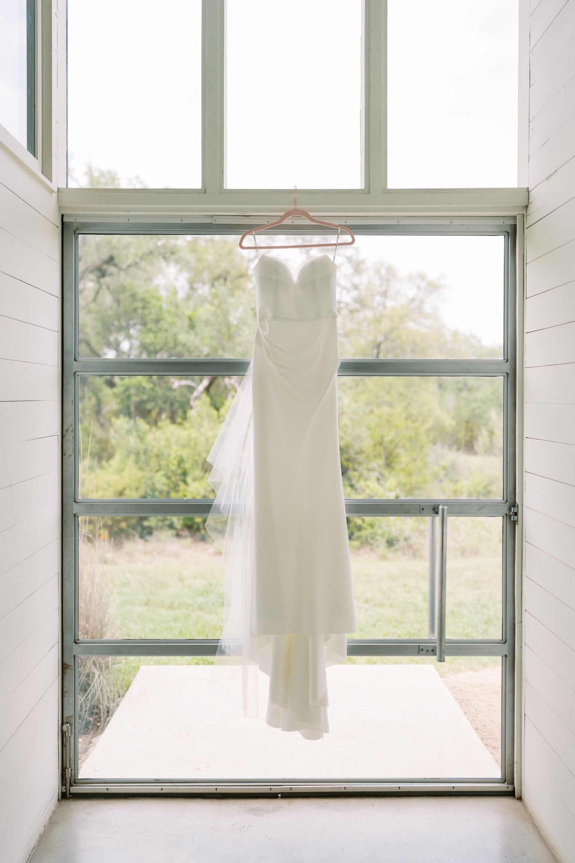 Wedding dress hanging on a pink hanger in front of a window, overlooking a green landscape.