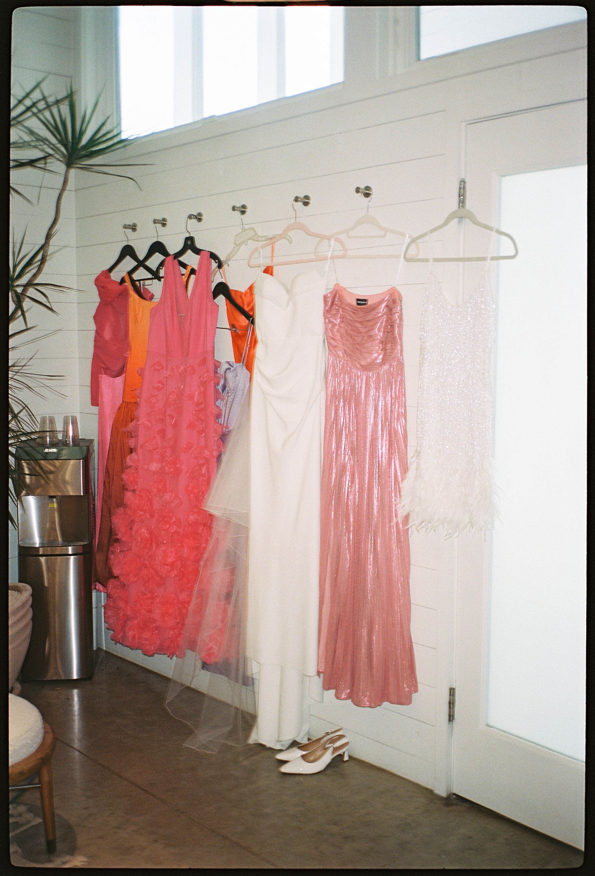 Dresses hanging on a white wall, including pink, orange, and sequined gowns. A room with a doorway.